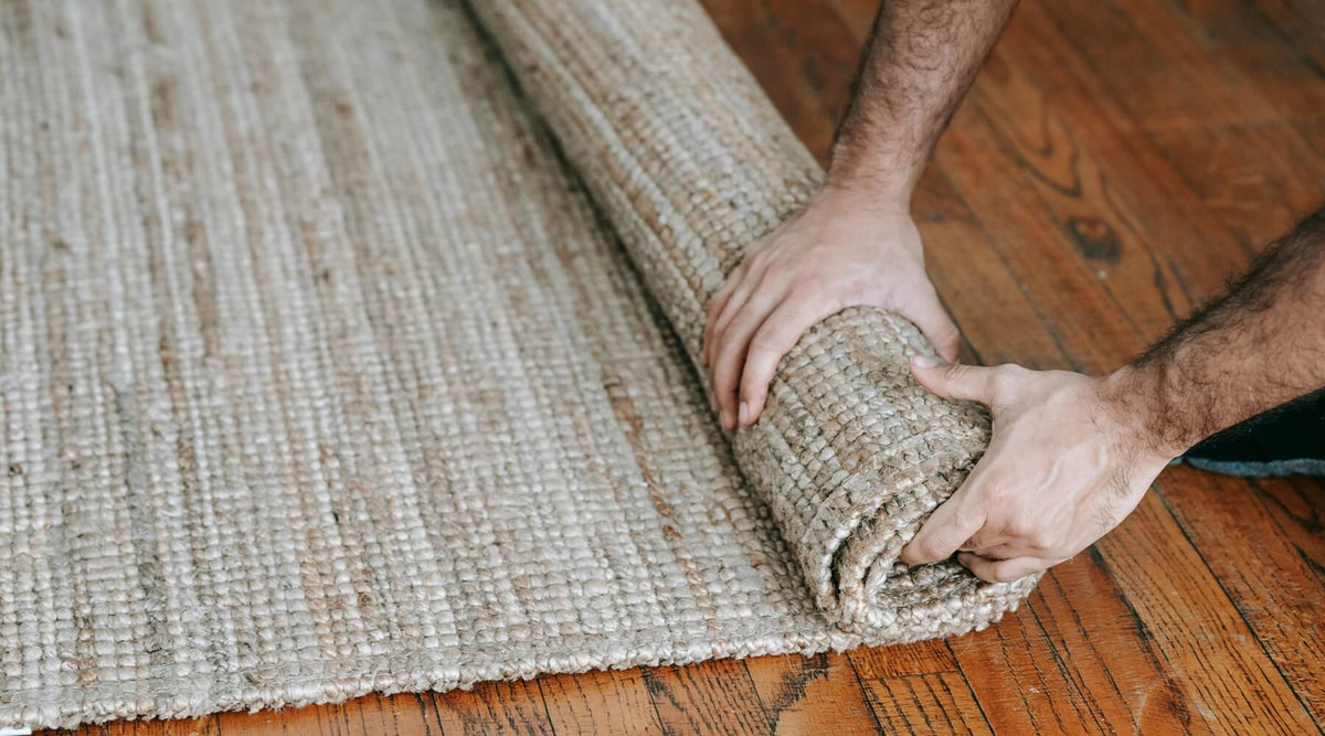 A person's hands are shown rolling up a light brown, textured area rug on a wooden floor, which could be part of Carpet Tips and Tricks for Cat Owners