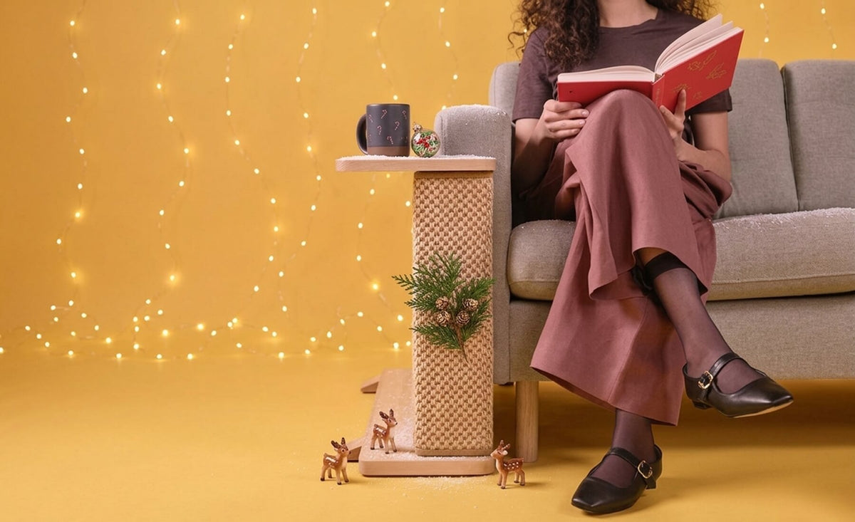 Cat-e-Corner as a Christmas gift styled on a sofa armrest with a holiday mug, pine sprig, and string lights